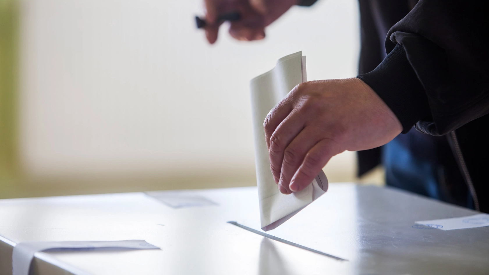 Jemand steckt mit der linken Hand einen gefalteten Wahlzettel in ein Wahlurne.